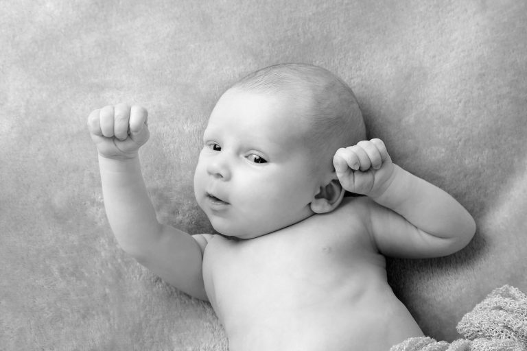 Black and white photo of a baby lying on a soft blanket with both arms raised and fists closed, looking slightly upwards—perfect for timeless newborn photos Edinburgh families will cherish.