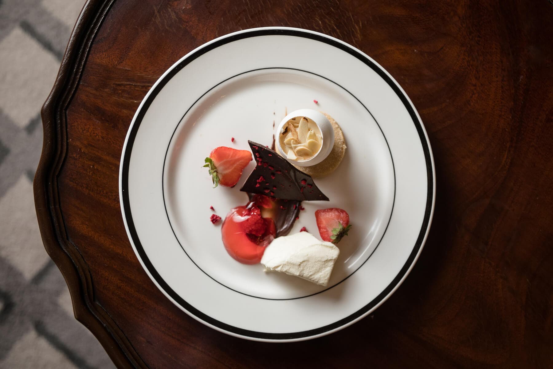A plated dessert at Mansfield Traquair Wedding Venue features a white meringue, chocolate shard, strawberries, a dollop of cream, and a small tart, beautifully arranged on a white plate with a dark wooden table background.
