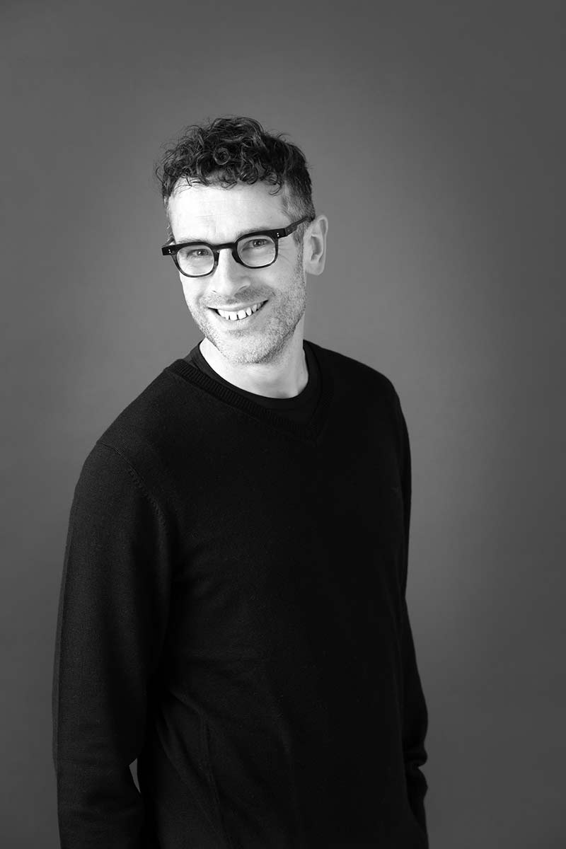 Niels wearing a dark jumper, standing and smiling in front of a plain background. Black and white photo 