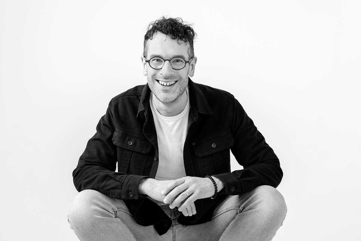 Niels wearing a dark jacket and light jeans, sits cross-legged and smiles at the camera against a plain background.