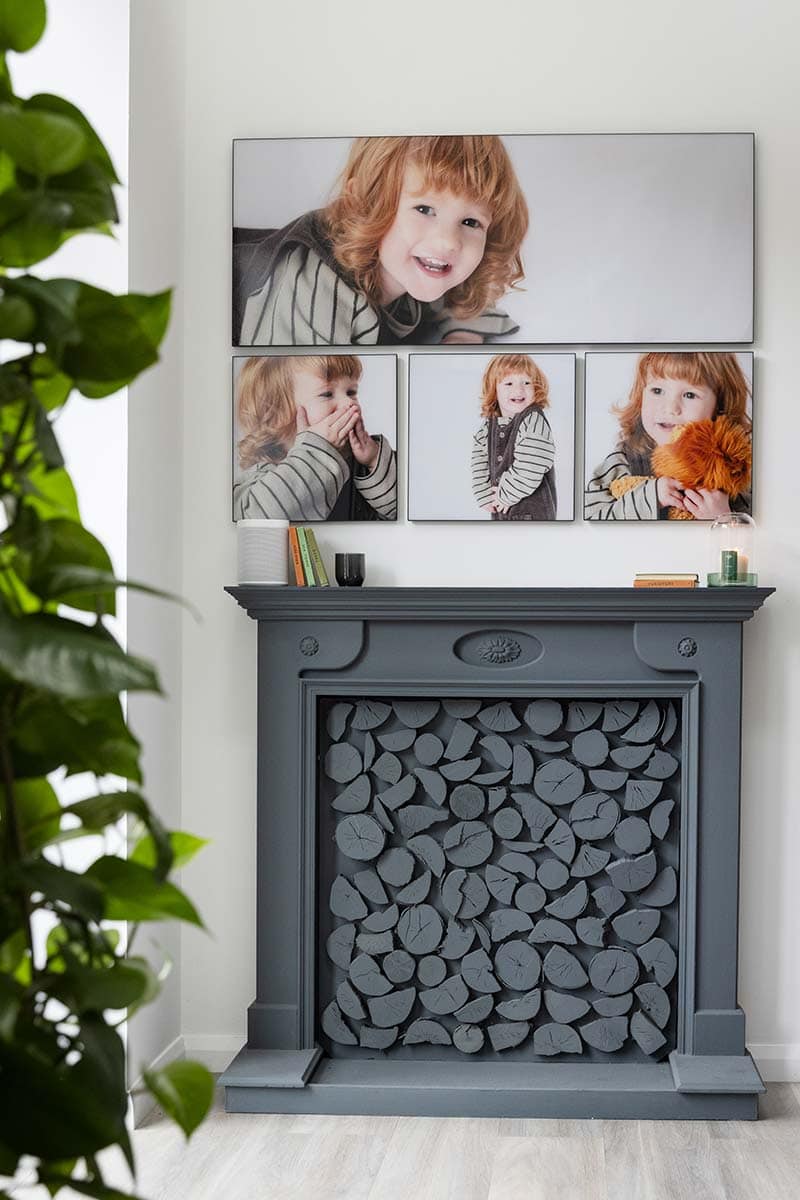 A grey decorative fireplace filled with painted wooden logs sits below three framed photos of a young child with light brown hair, set against a white wall, evoking the airy charm of blue sky photography.