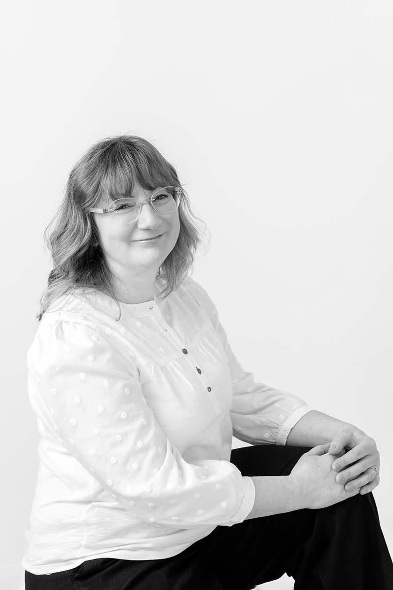 Bianca sits with one knee up, wearing a white blouse and dark trousers against a plain light background.