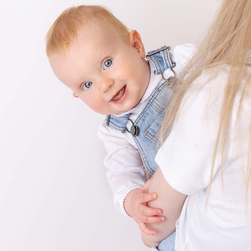 A baby with fair hair and blue eyes, dressed in denim dungarees and a white shirt, is held by an adult with long blonde hair, both facing a light background.