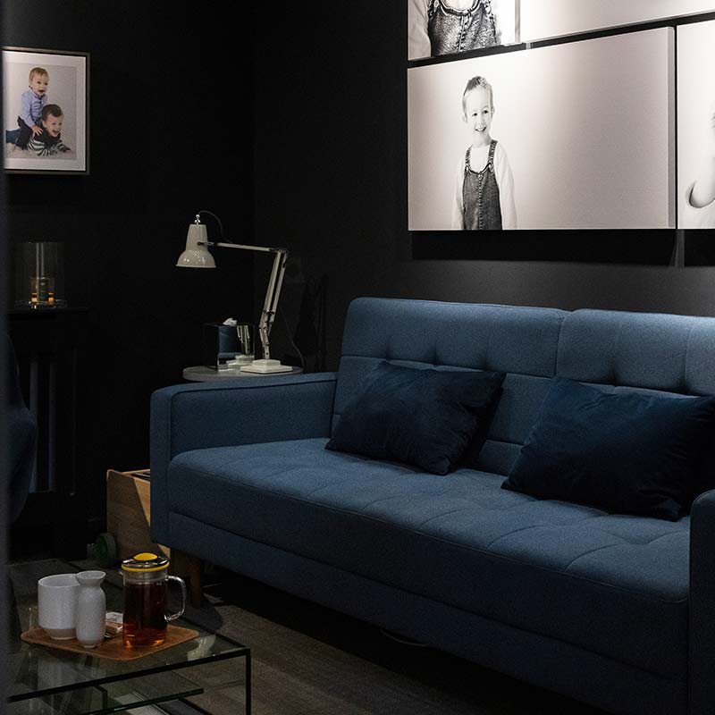 A dark studio view room with a blue sofa, two navy cushions, wall art of children by Edinburgh family photographers, a desk lamp, mugs, and a glass teapot on a glass coffee table.