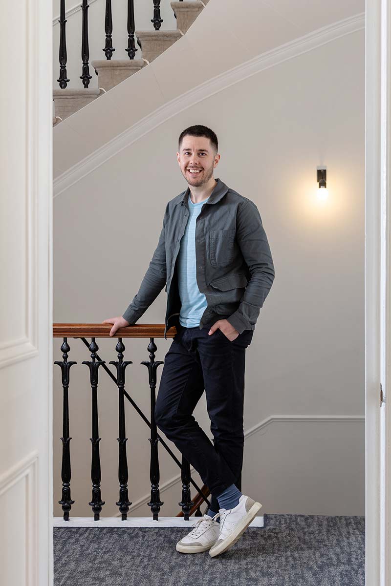 A man stands on an indoor staircase landing, leaning against a wooden bannister, dressed casually in a jacket, t-shirt, trousers, and trainers—an authentic moment of people photography in Edinburgh.
