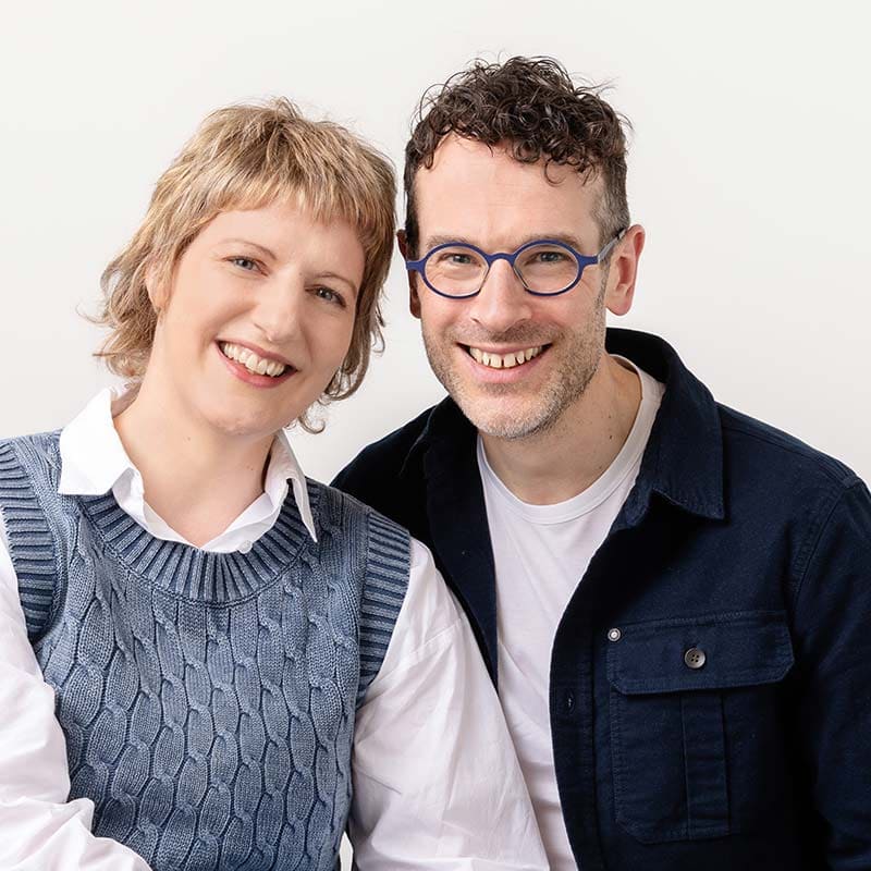 Niels and Alie Calvert smiling at the camera; one wears a white shirt with a blue jumper and the other wears glasses, a white shirt, and a dark jacket