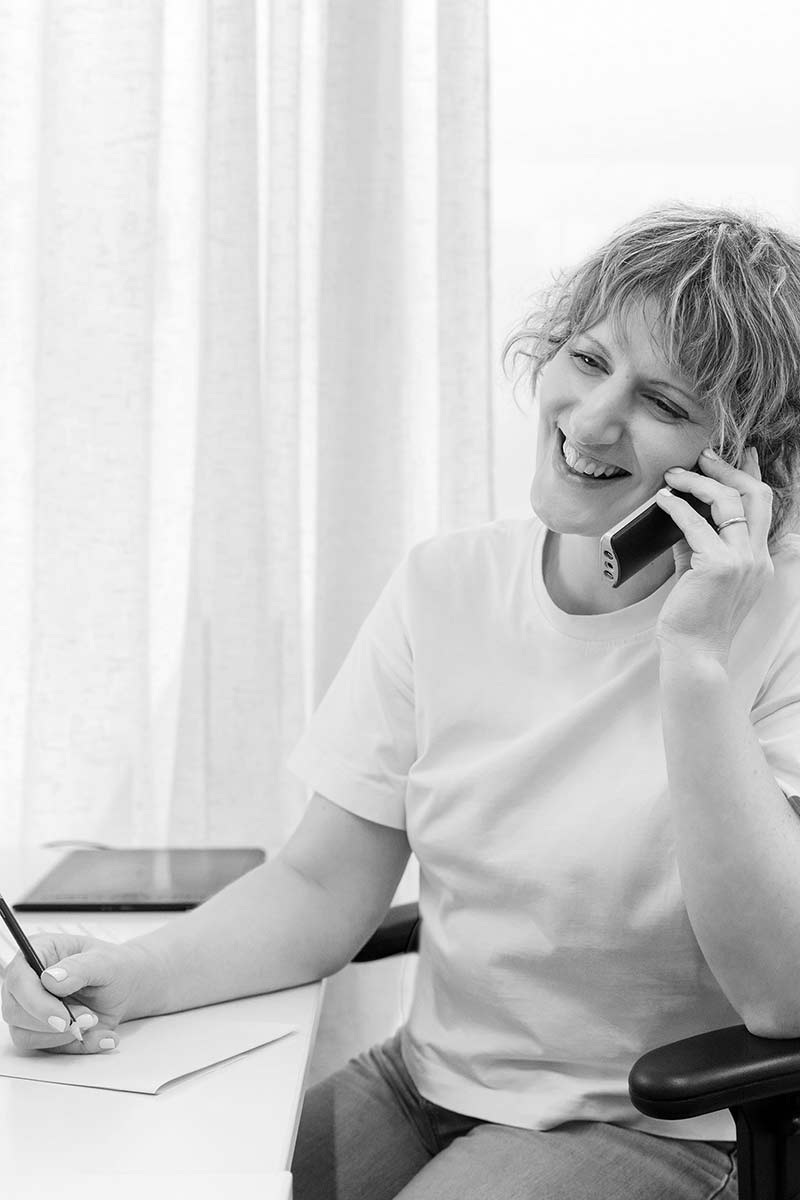 Photographer Alie Calvert sits at a desk, talking on a mobile phone and writing on a piece of paper, smiling