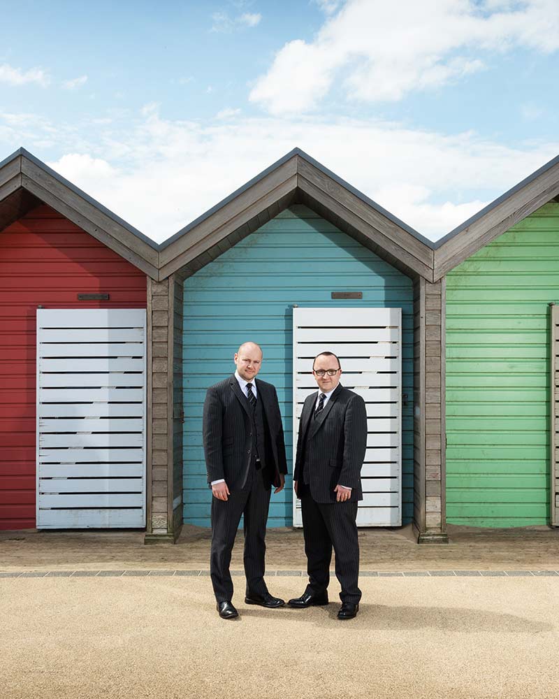 Two men in suits stand in front of colourful beach huts—one red, one blue, and one green—under a partly cloudy sky, capturing a vibrant scene ideal for Commercial Photography Edinburgh.