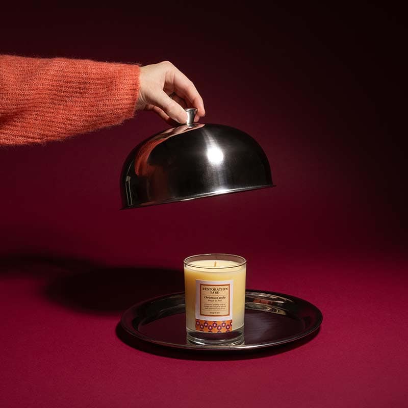 A hand lifts a silver cloche to reveal a scented candle in a glass jar on a matching silver tray, set against a red background—an elegant example of commercial photography Edinburgh style.