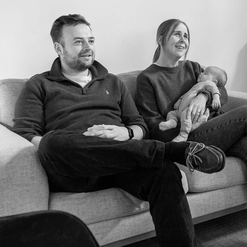 A man and a woman sit on a sofa; the woman cradles a baby in her arms. All three face forward, appearing relaxed. The black and white scene captures a serene moment perfect for newborn photography.