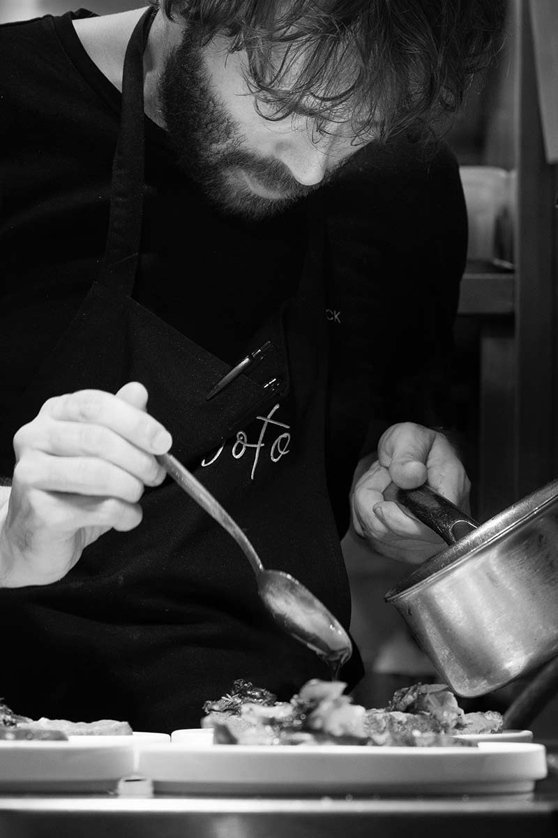 A chef wearing an apron is carefully plating food, pouring sauce from a small pan and using a spoon to add detail to the dishes. The image is BW