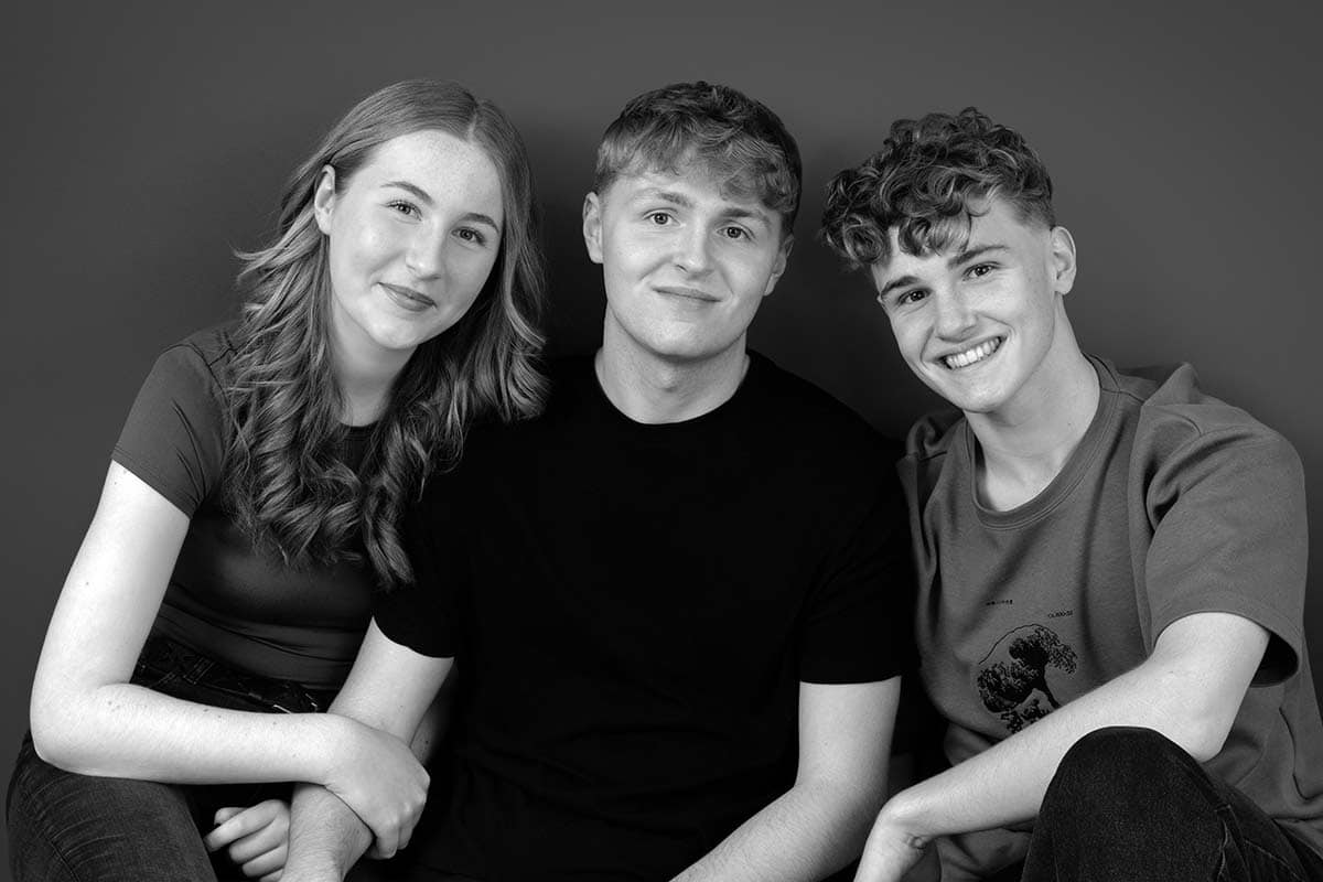 Three young people sit closely together in front of a plain studio background, smiling at the camera. The image is black and white.